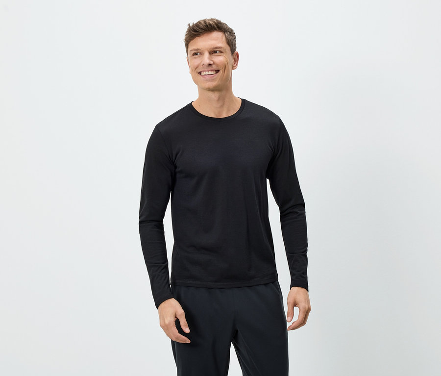 Ein lächelnder Mann trägt ein schwarzes Langarmshirt und eine schwarze Hose vor einem weißen Hintergrund.