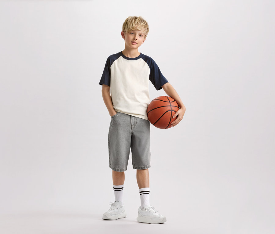 Ein Junge mit blonden Haaren hält einen Basketball in der Hand, trägt ein helles T-Shirt mit marineblauen Ärmeln, graue Shorts und weiße Socken mit schwarzen Streifen.