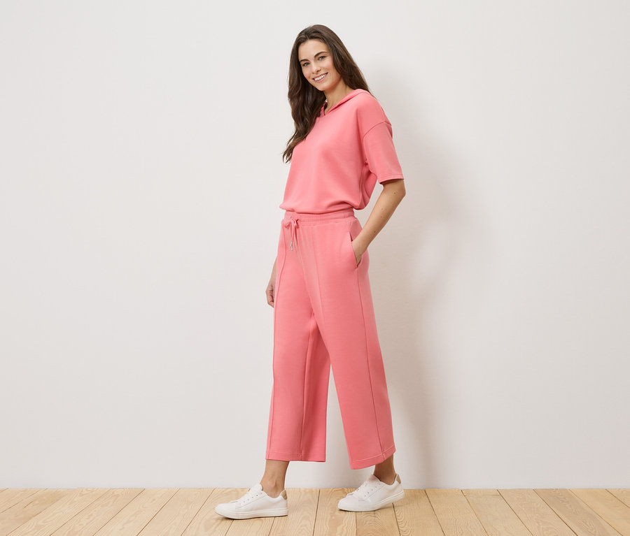 Frau trägt einen korallenroten Trainingsanzug mit Kapuzenoberteil und Culottes sowie weiße Turnschuhe.