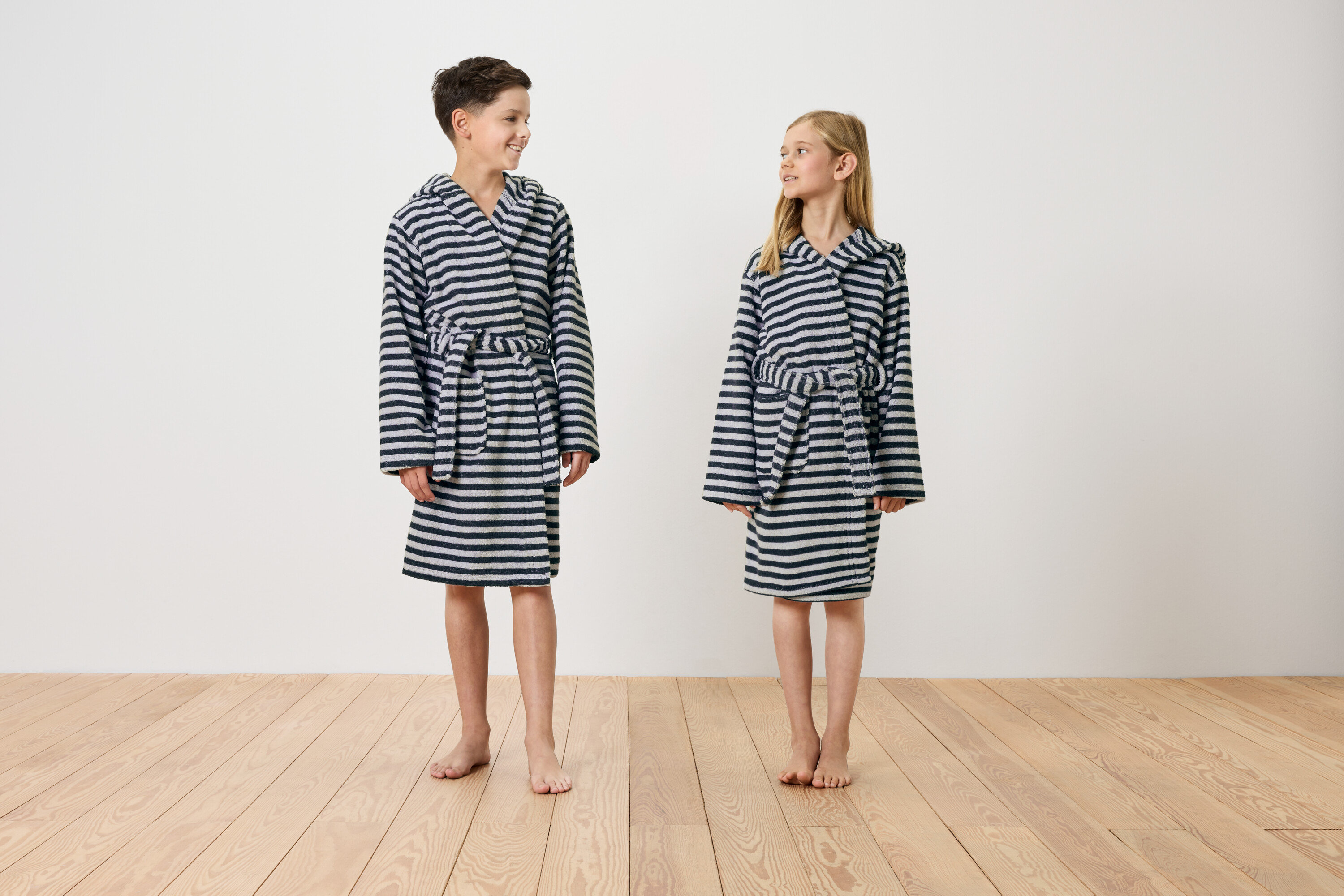 Ein Junge und ein Mädchen stehen barfuß auf einem Holzboden und tragen gestreifte Bademäntel mit Kapuze.