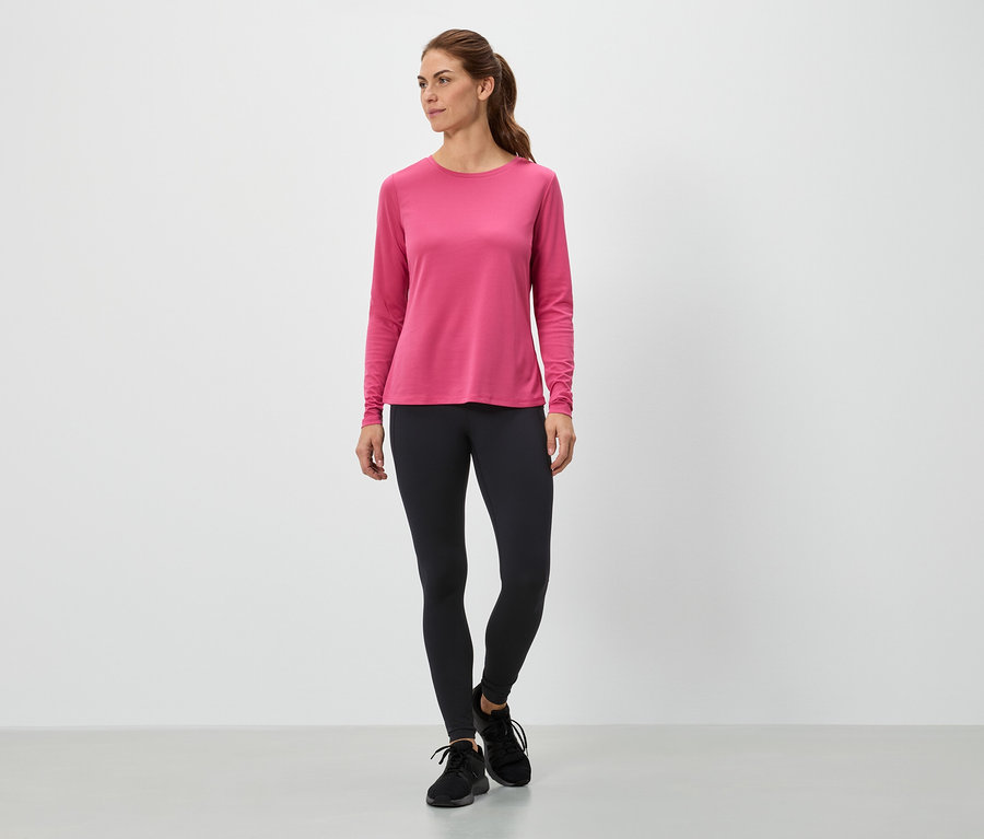 Frau steht in einem pinkfarbenen Langarmshirt und schwarzen Leggings.