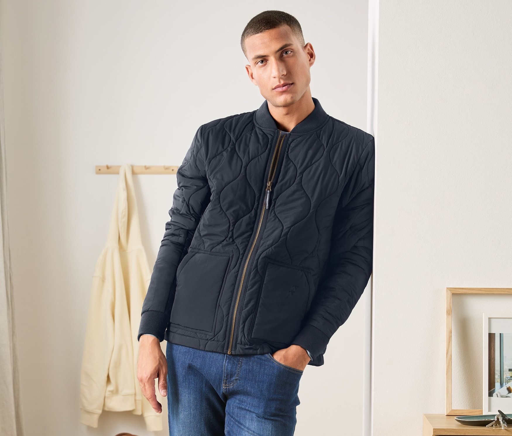 Mann lehnt an Wand, trägt blaue Steppjacke, Jeans und weiße Turnschuhe.