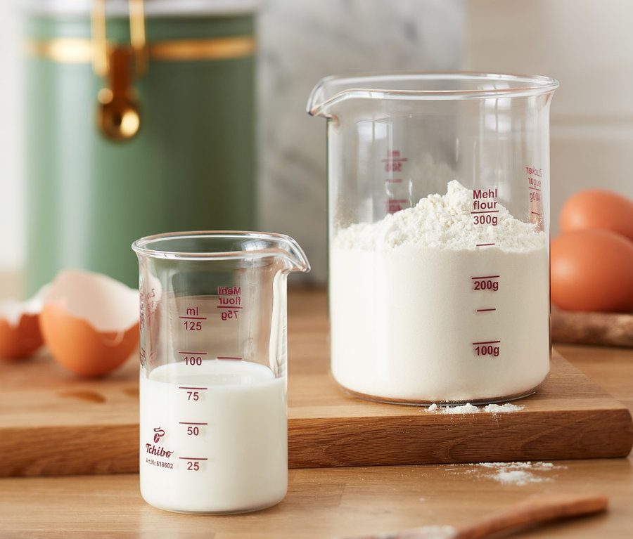 Zwei Messbecher, einer mit Milch und einer mit Mehl, stehen auf einem Holztisch neben Eiern.