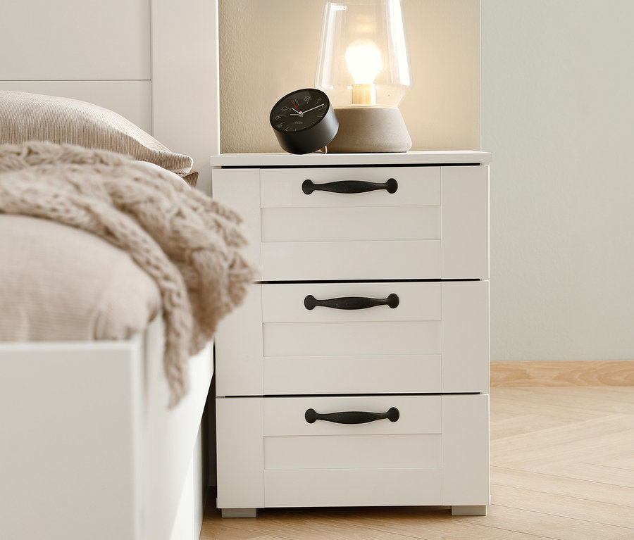Weißer Nachttisch »Cottage« mit drei Schubladen, auf dem eine Lampe und ein Wecker stehen.