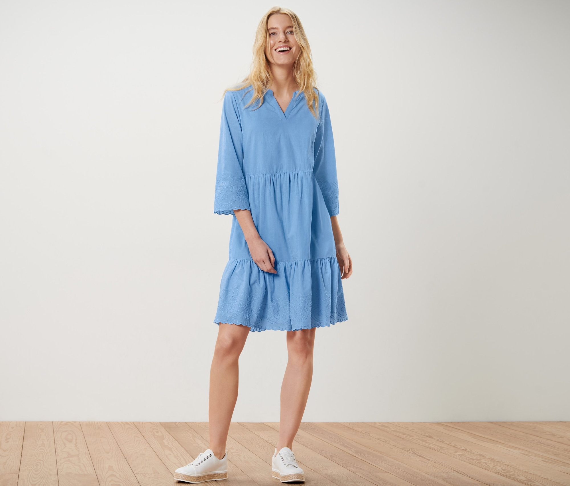 Eine blonde Frau steht und lächelt, sie trägt ein blaues Webkleid mit Stickerei und weiße Turnschuhe.