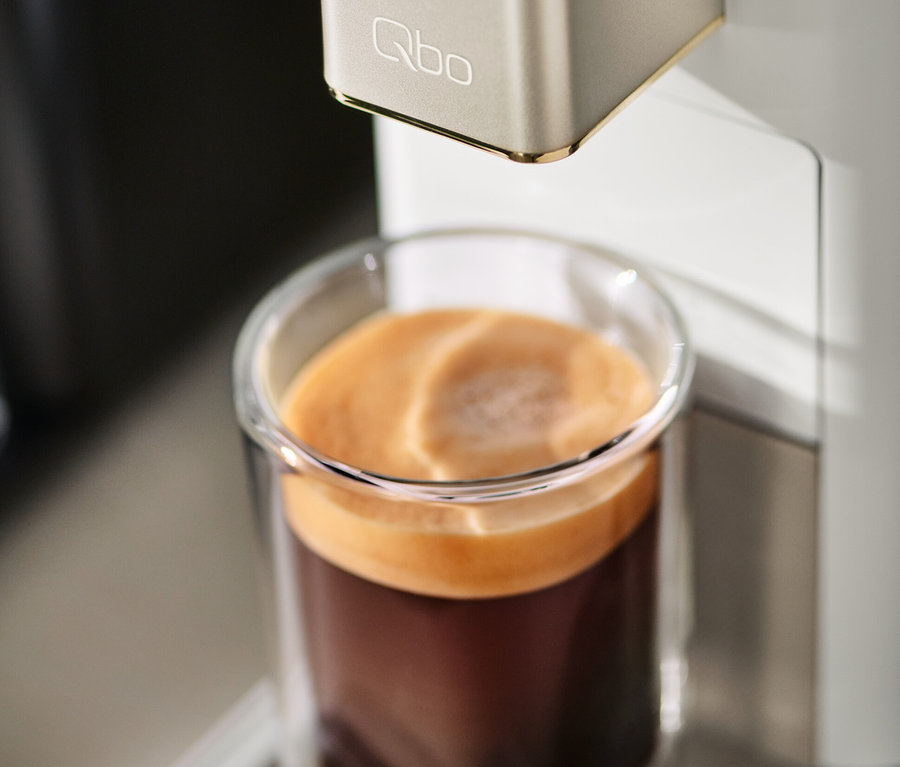 Qbo ICONIC Light Gold Kaffeemaschine befüllt ein Glas mit Kaffee.