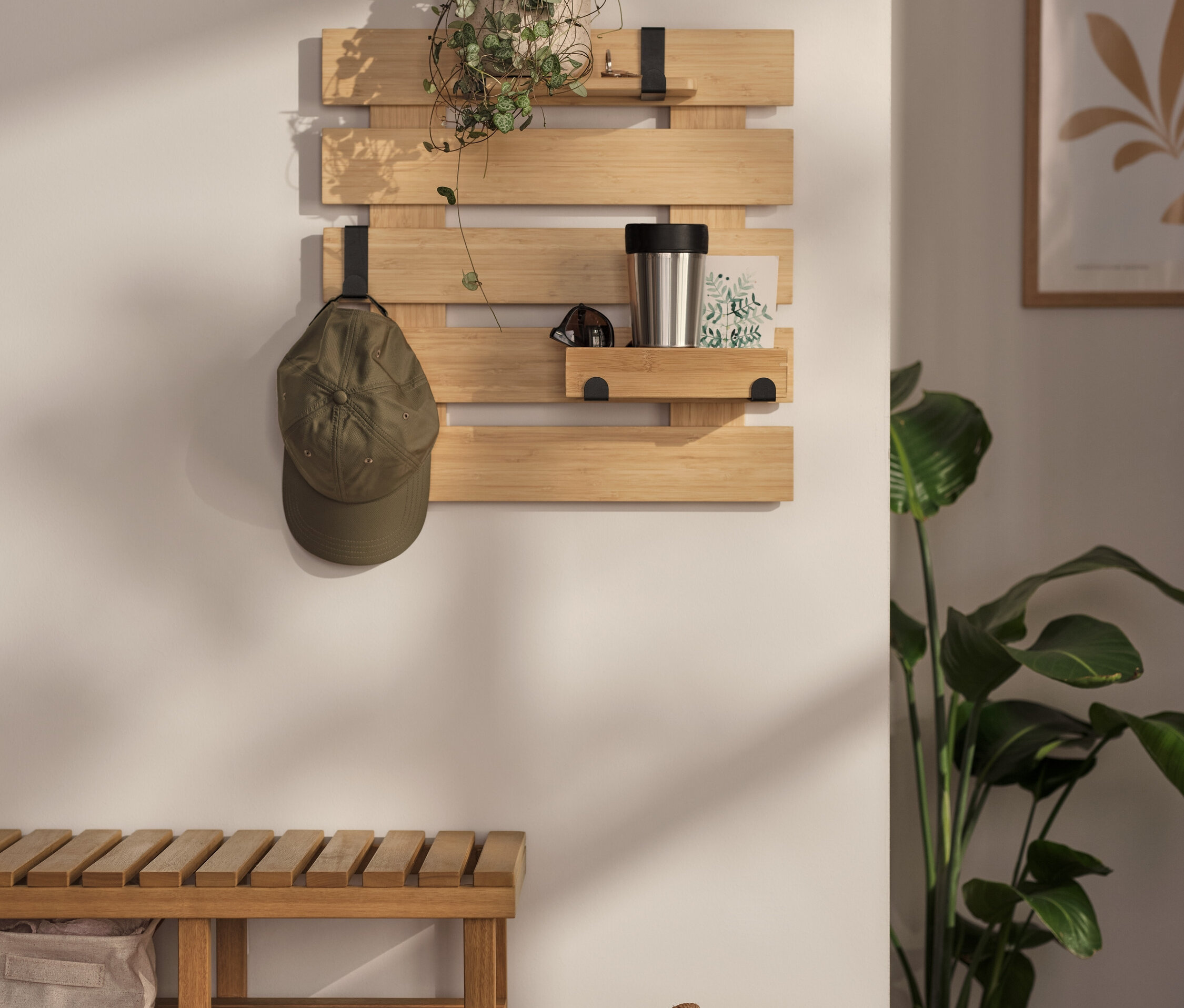 An der Wand hängt ein Wandregal mit flexiblen Haken und einem Boden. Darauf befinden sich eine Pflanze, eine Thermoskanne und eine Karte. An einem der Haken hängt eine Kappe.