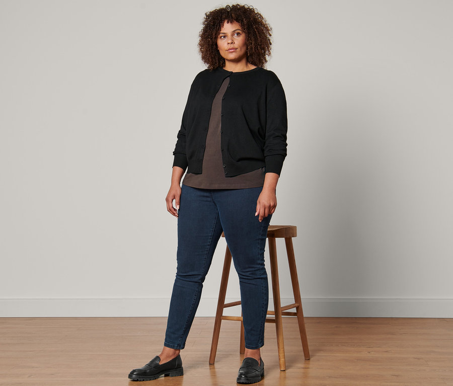 Frau steht und trägt einen schwarzen Feinstrick-Cardigan, ein Shirt mit 3/4-Arm, Treggings und Leder-Loafer.