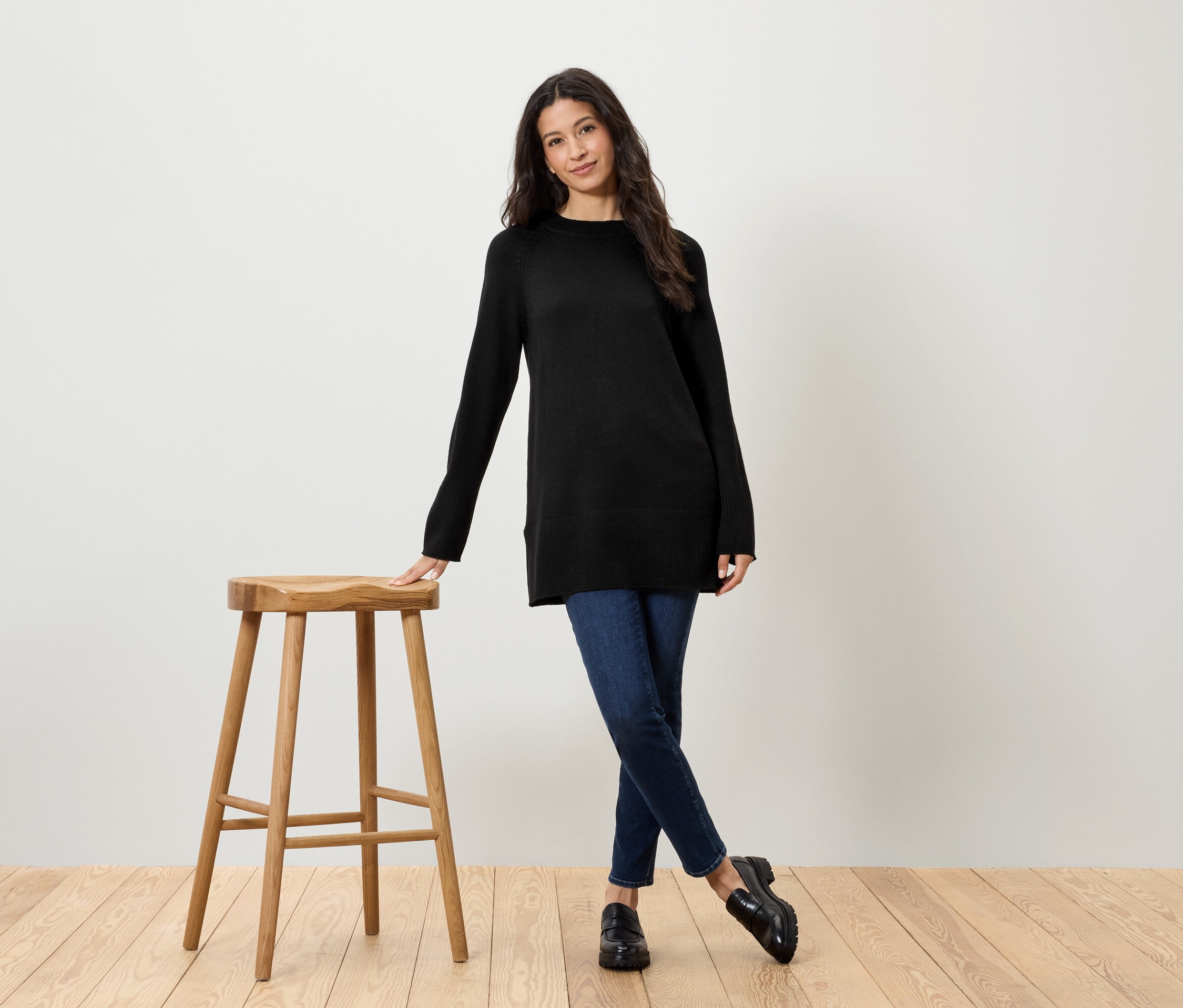 Frau mit schwarzem Pullover und Jeans steht neben einem Holzhocker.