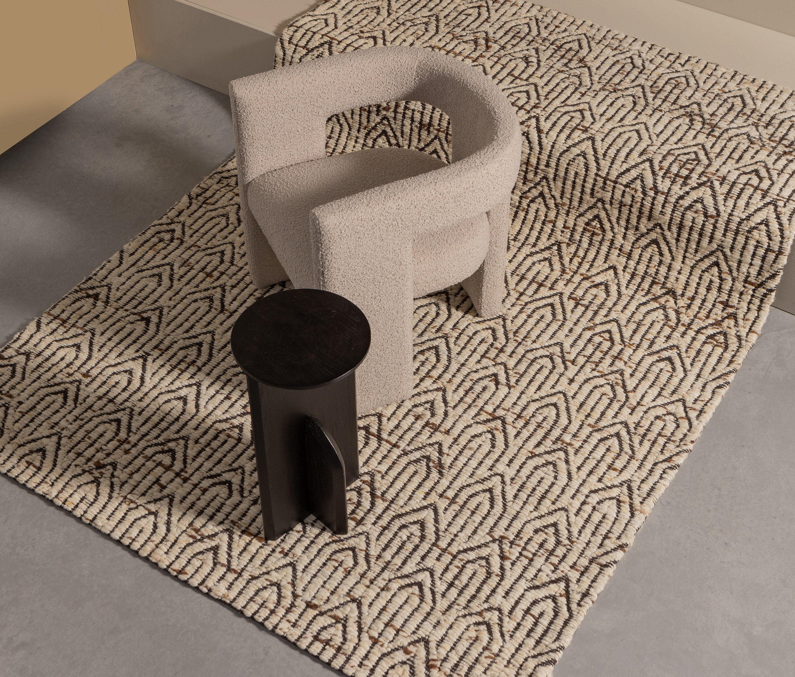 Ein cremefarbener Sessel und ein schwarzer Hocker stehen auf dem beige Woood Teppich Joslin.