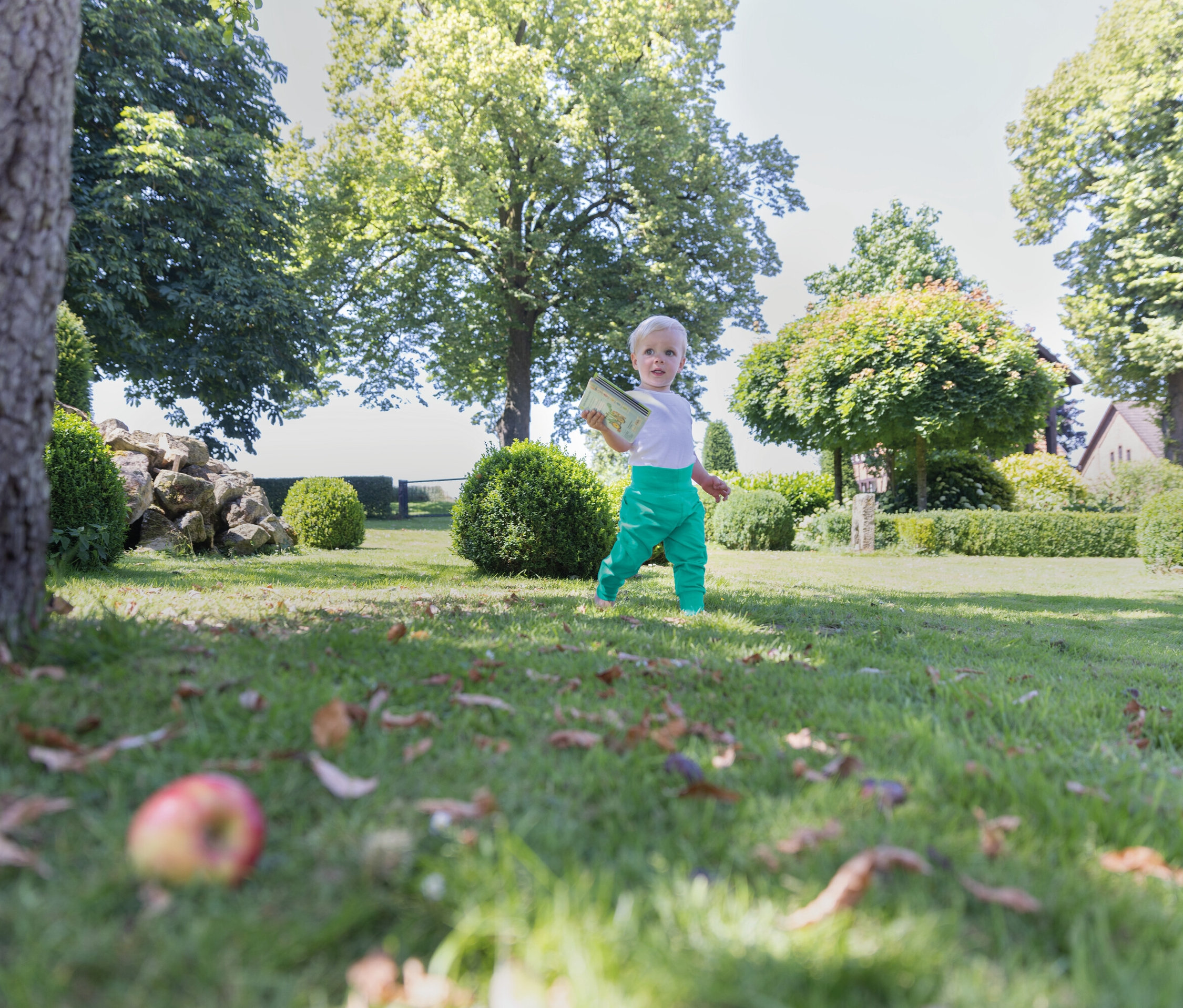 Ein Kleinkind in grüner Hose geht auf einer Wiese in einem Garten.