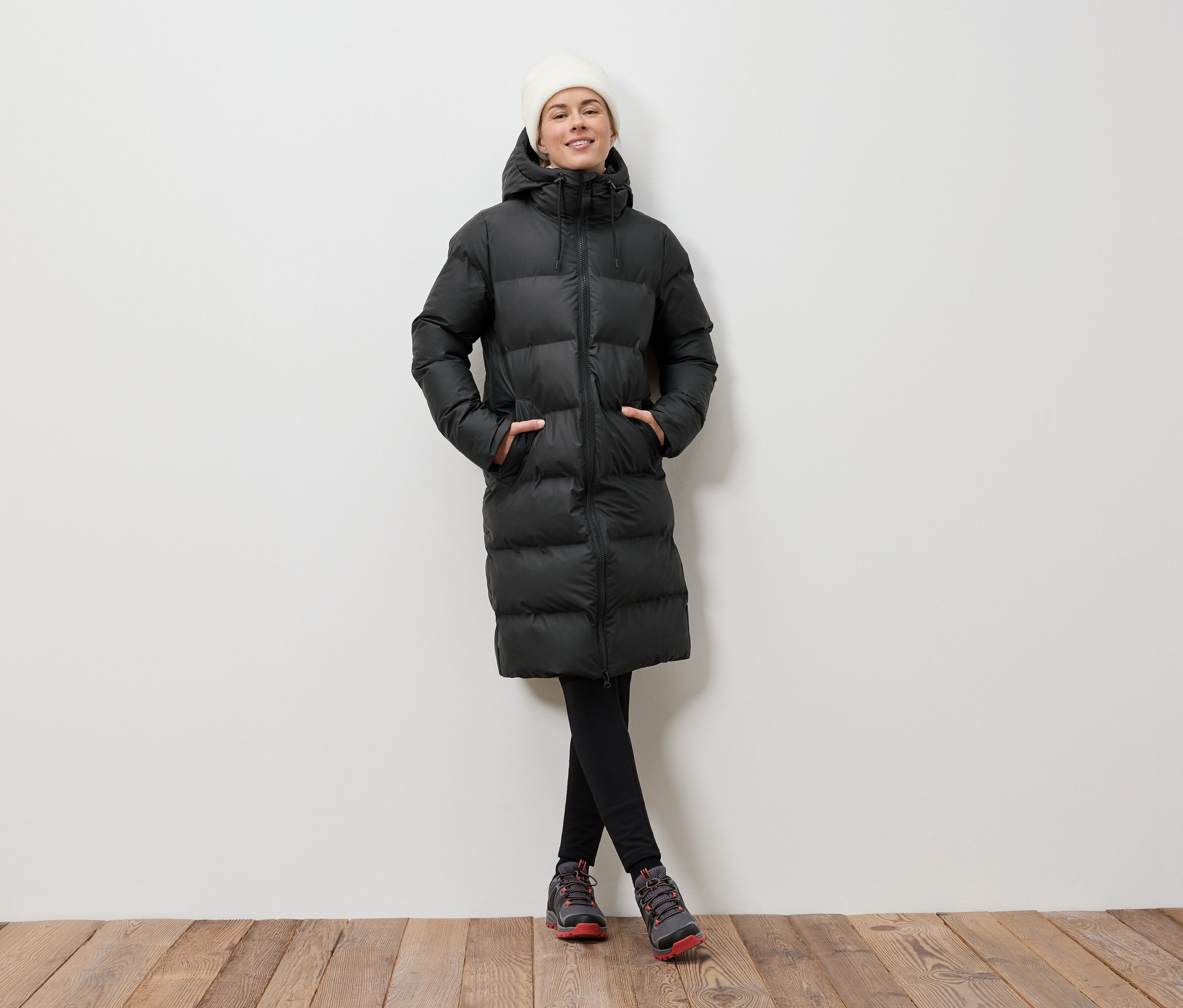 Frau posiert auf einem Holzboden in einer schwarzen Daunenjacke und einer weißen Mütze.