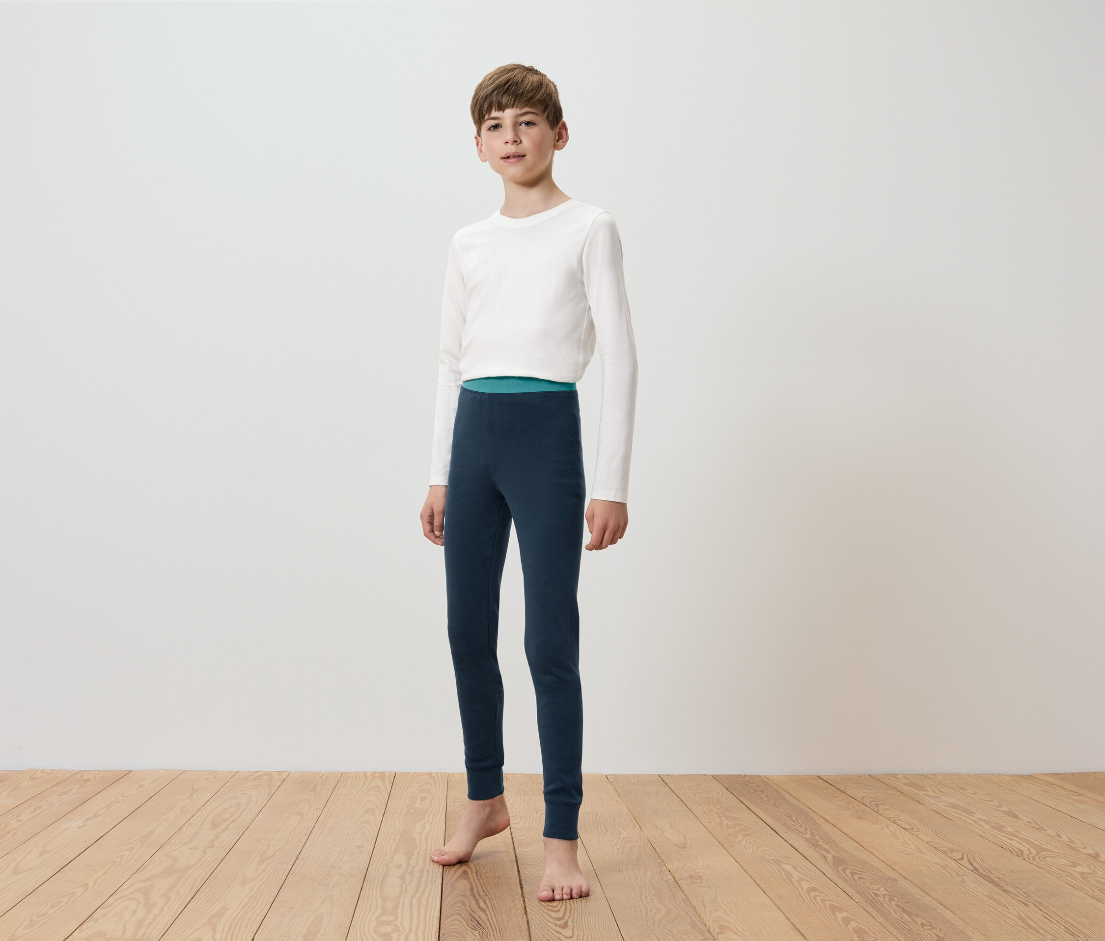 Junge mit weißem Langarmshirt und dunkelblauer Leggings steht barfuß auf Holzboden vor einer weißen Wand.
