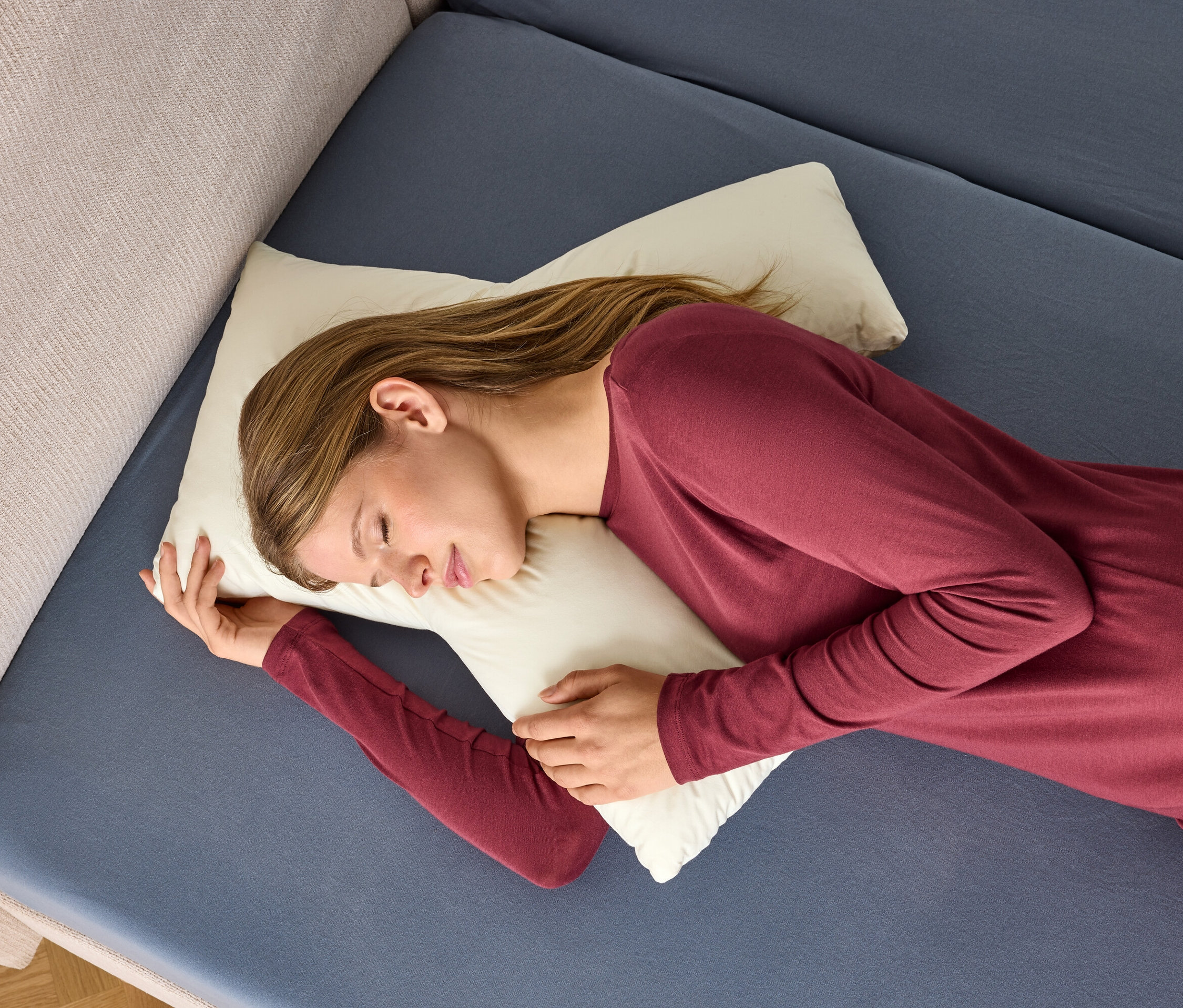 Frau im bordeauxroten Nachthemd schläft auf einem weißen Kissen auf einem blauen Bett.