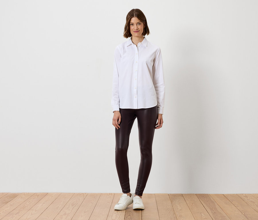 Frau im weißen Hemd und braunen Leggings steht auf Holzboden.