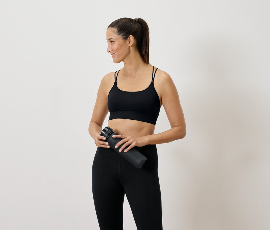 Frau in schwarzer Sportbekleidung hält eine Wasserflasche in der Hand und schaut zur Seite.