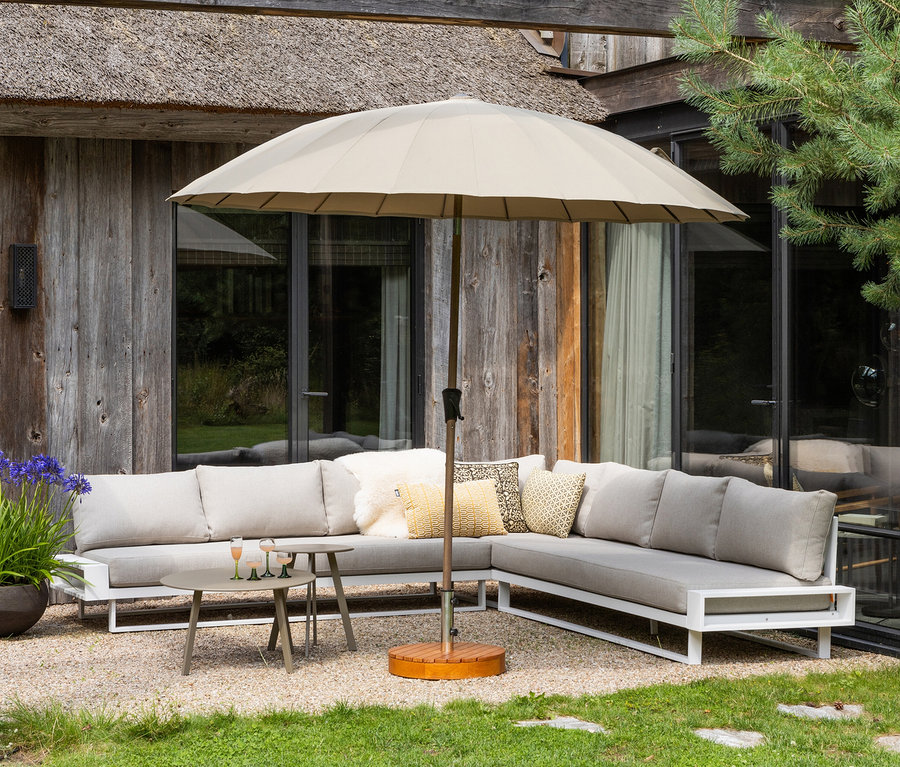 Ein graues Ecksofa mit weißen Rahmen steht auf einem Kiesweg. Ein beiger Sonnenschirm steht daneben.