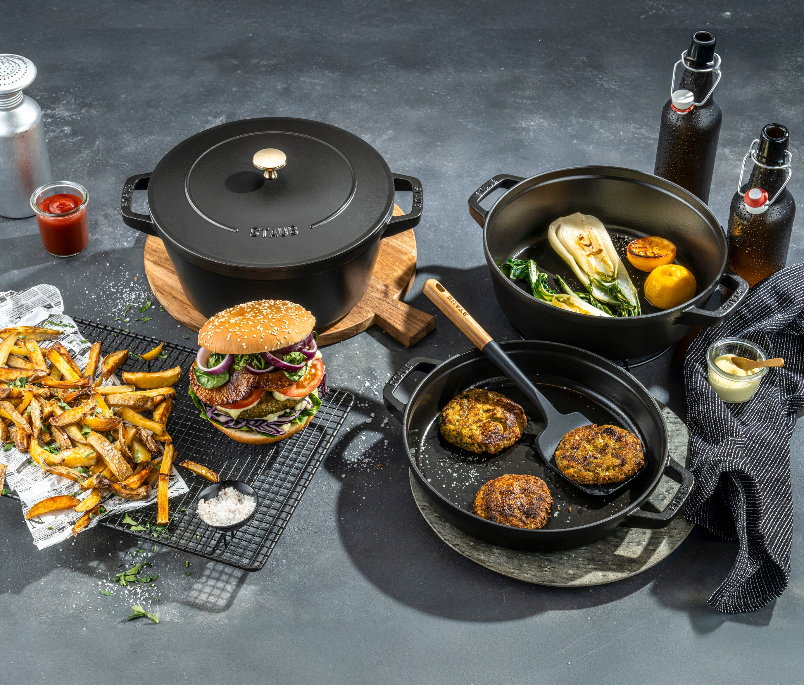 Verschiedene Speisen, darunter ein Burger, Pommes und Gemüse, werden zusammen mit einem STAUB Kochtopf-Set »La Cocotte«, 3-teilig, präsentiert.