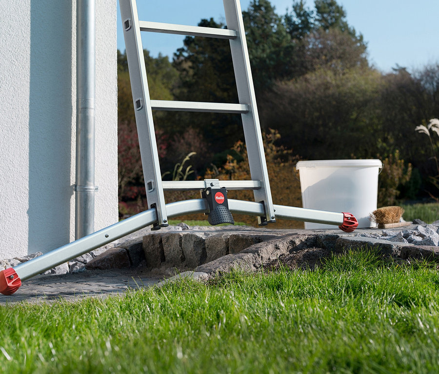 Eine silberne Leiter steht auf einem steinigen Untergrund vor einer weißen Hauswand, neben einem weißen Eimer und einer Bürste.