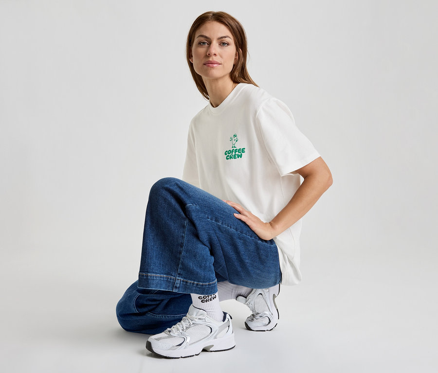 Frau hockt in Jeans und T-Shirt mit dem Aufdruck "Coffee Crew" und trägt weiße Turnschuhe.