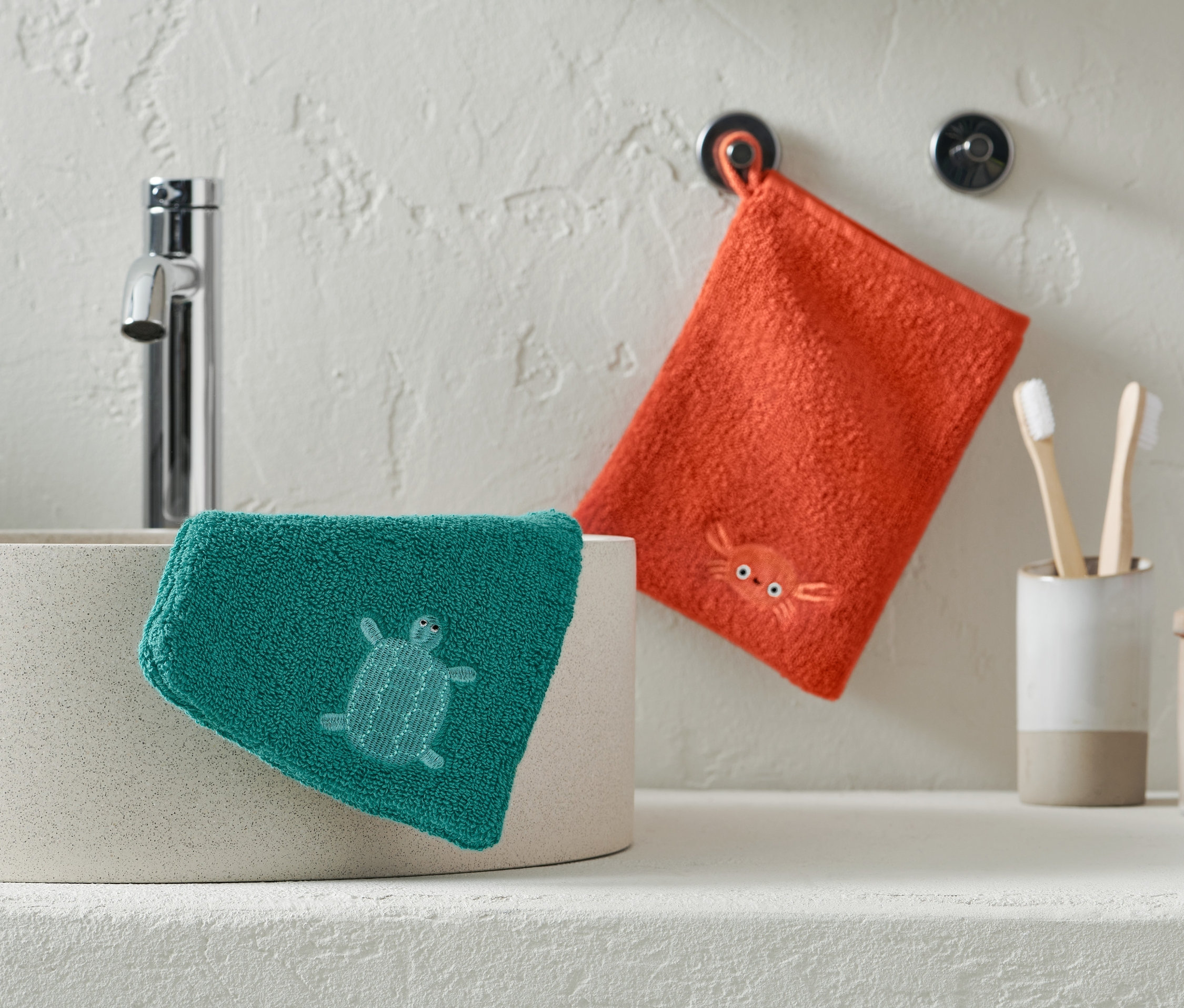 Ein grüner Waschlappen mit einer Schildkröte liegt auf einem Waschbecken, ein orangefarbener Waschlappen mit einer Krabbe hängt an der Wand.