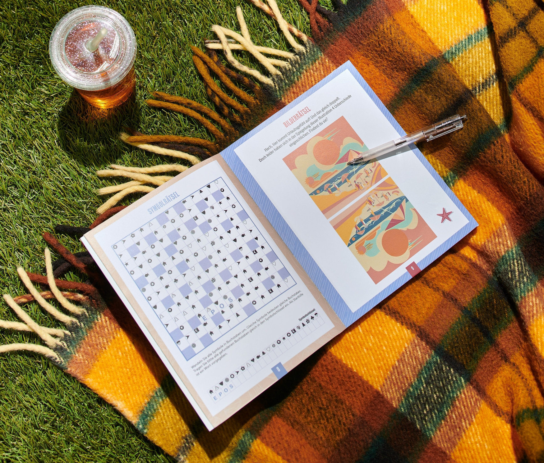 Auf einer Decke mit Fransen im Gras liegt ein geöffnetes Buch »Gute-Laune Sommer-Rätsel« und daneben ein Glas mit Getränk.