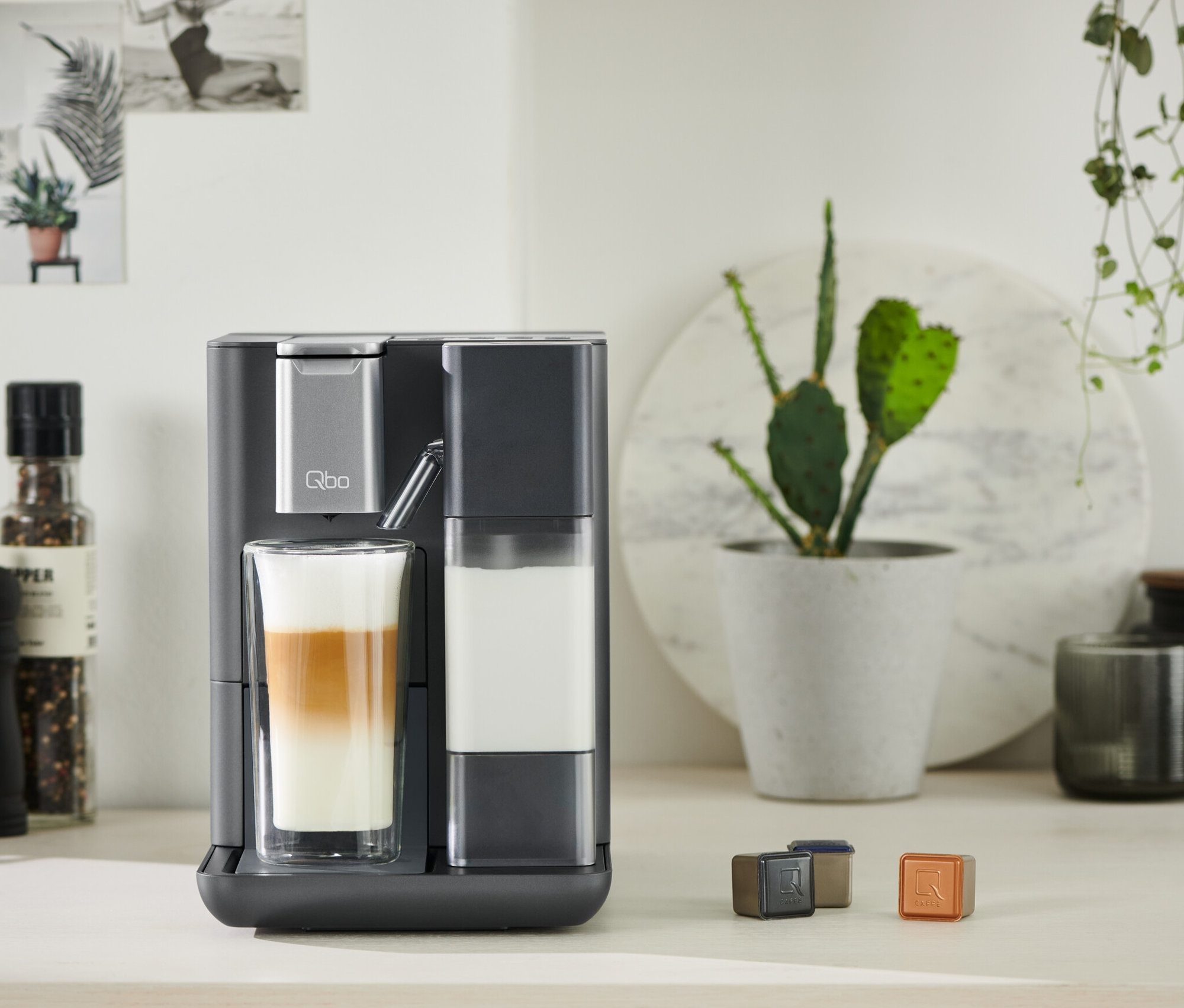 Graue Qbo MILK MASTER bereitet Getränk in Glas zu. Kapseln liegen auf Tisch.