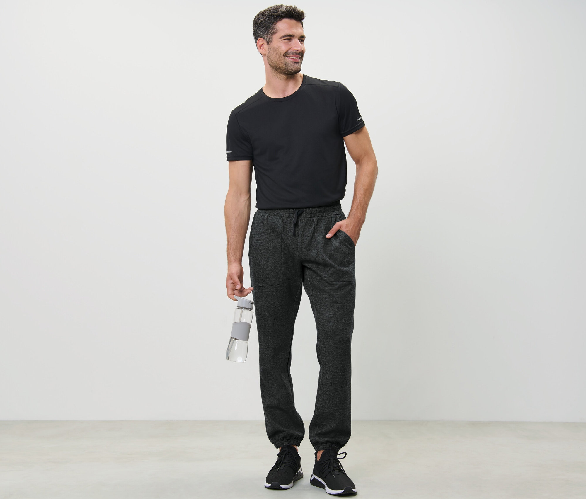 Mann im schwarzen T-Shirt hält eine Wasserflasche und trägt eine Sweathose.