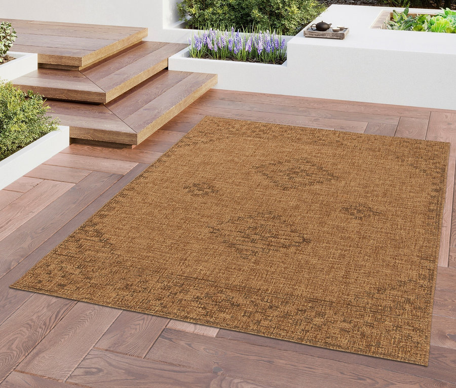 Ein brauner Teppich »Roy« liegt auf einer Holzterrasse. Im Hintergrund sind Treppen, Pflanzen und ein Haus zu sehen.