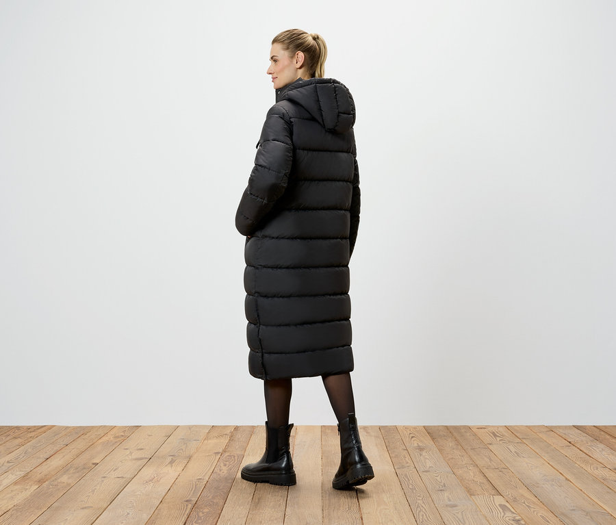 Eine Frau steht mit dem Rücken zur Kamera und trägt einen langen, schwarzen Mantel mit Kapuze, eine schwarze Strumpfhose und schwarze Stiefel.