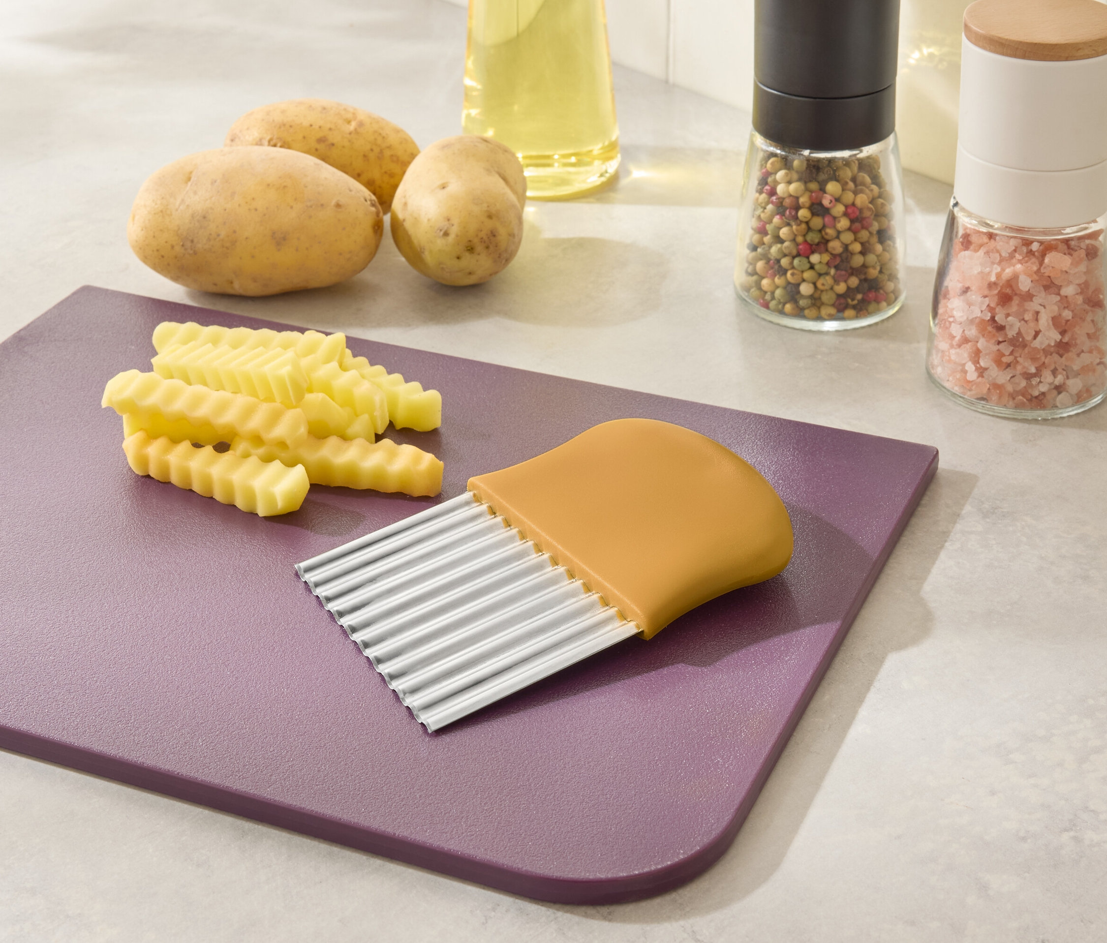 Auf einem lila Schneidebrett liegen geschnittene Pommes Frites und ein gelber Pommesschneider. Im Hintergrund befinden sich Kartoffeln, Öl und Gewürze.