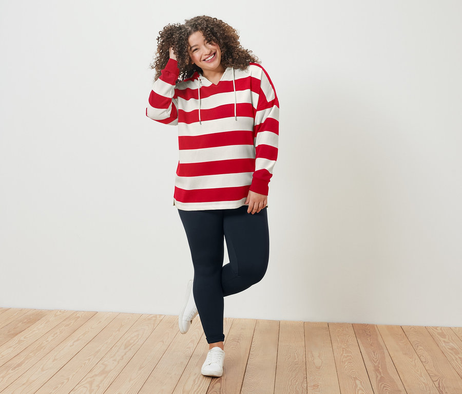 Frau mit lockigem Haar, rot-weiß gestreiftem Oberteil und blauen Leggings steht auf einem Holzboden.