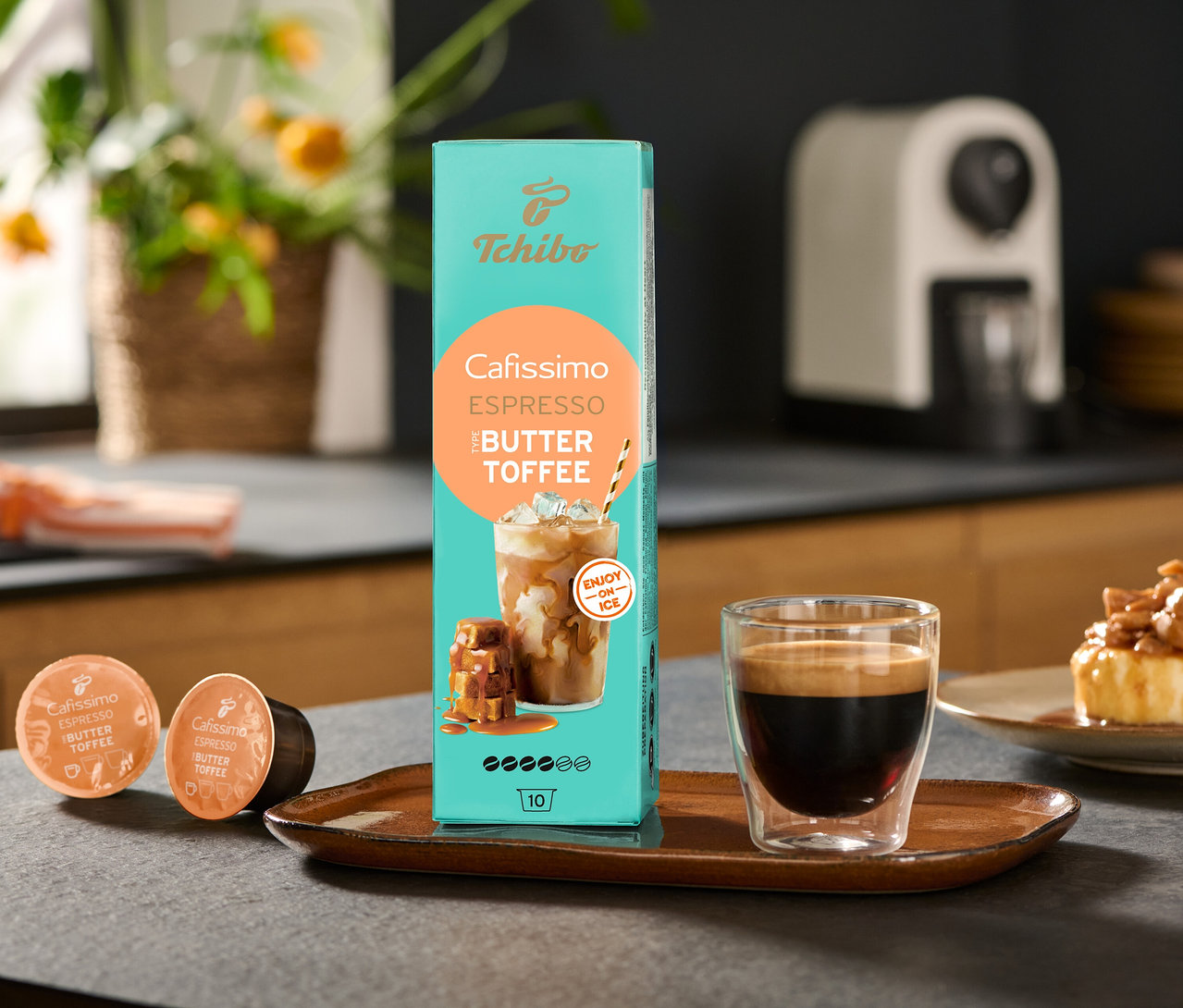Eine Packung Cafissimo Flavoured Espresso – Buttertoffee und eine Cafissimo pure+, white Kaffeemaschine stehen auf einem Tisch, zusammen mit einer Tasse Kaffee.