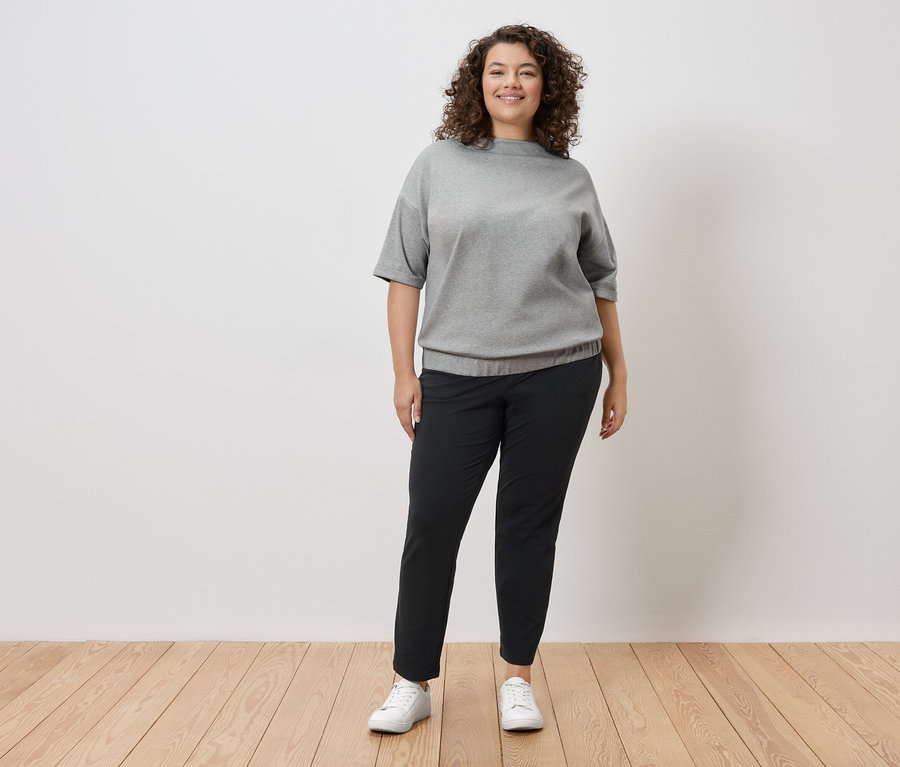 Eine Frau mit lockigem Haar steht vor einer weißen Wand und trägt ein graues Sweatshirt, eine schwarze Hose und weiße Turnschuhe.