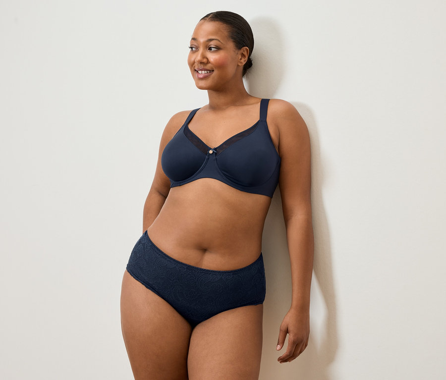 Frau mit marineblauem BH und Slip steht an einer weißen Wand und lächelt.