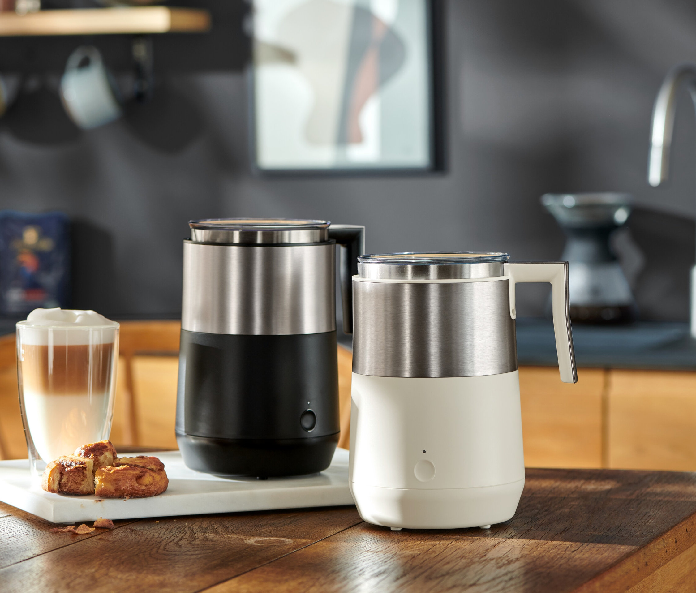 Zwei Induktions-Milchaufschäumer, einer schwarz, einer cremeweiß, stehen auf einem Holztisch neben einem Latte Macchiato Glas und Gebäck.