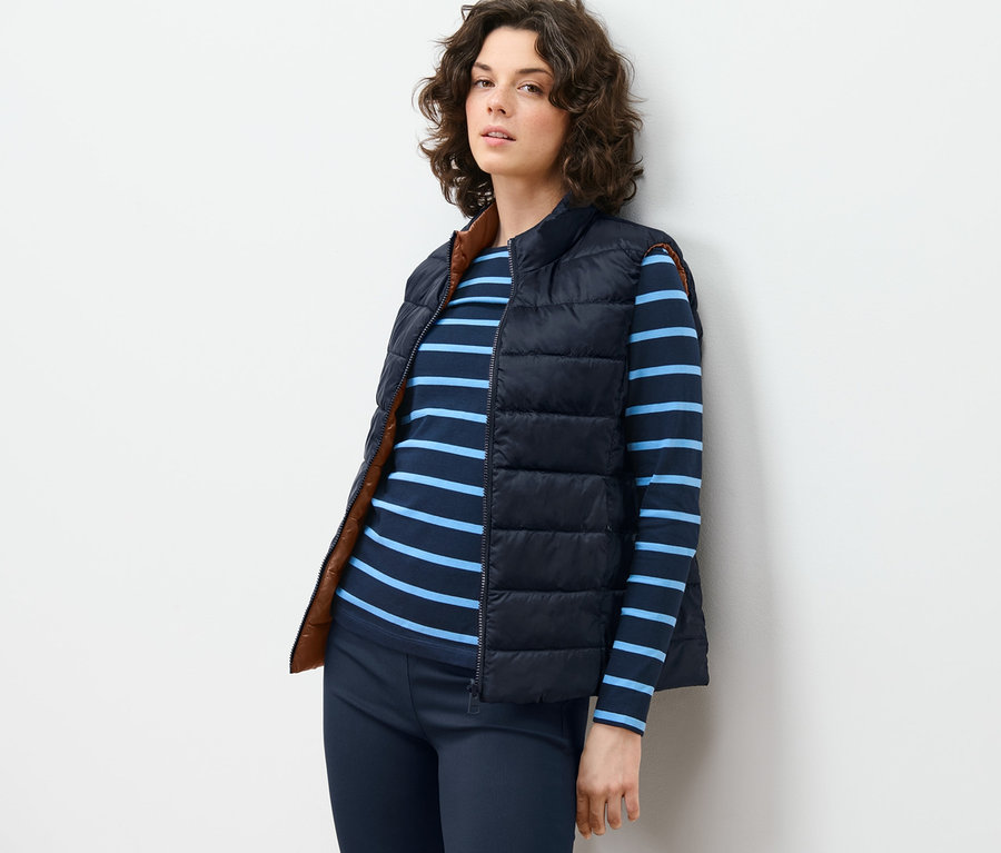 Frau lehnt an Wand, trägt Wende-Steppweste, gestreiftes Shirt und Hose.