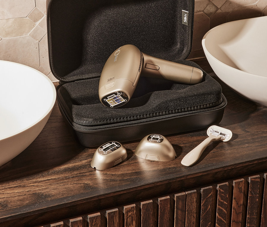 Goldenes Silk'n IPL Haarentfernungsgerät »Silk'n 7« in geöffnetem, schwarzen Etui mit Aufsätzen und Rasierer auf Holzoberfläche.