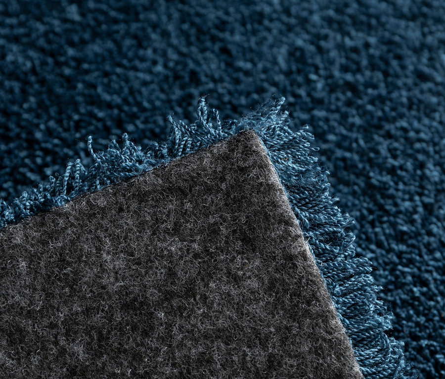 Detailansicht eines blauen Teppichs »Mellow« mit Fransen und grauer Unterseite.