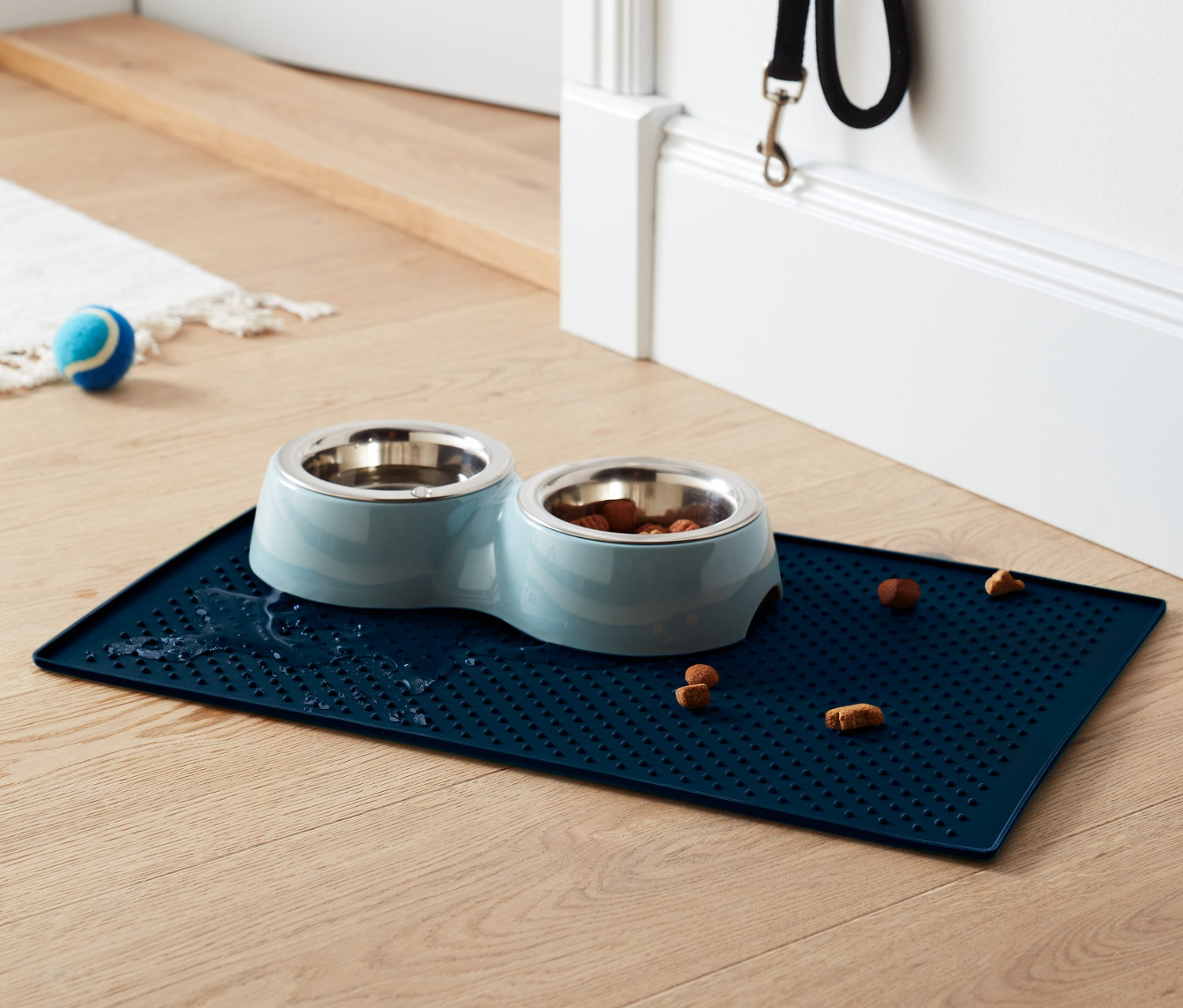 Blauer Futternapf-Untersetzer mit zwei Edelstahlnäpfen, einer mit Futter, auf Holzboden.