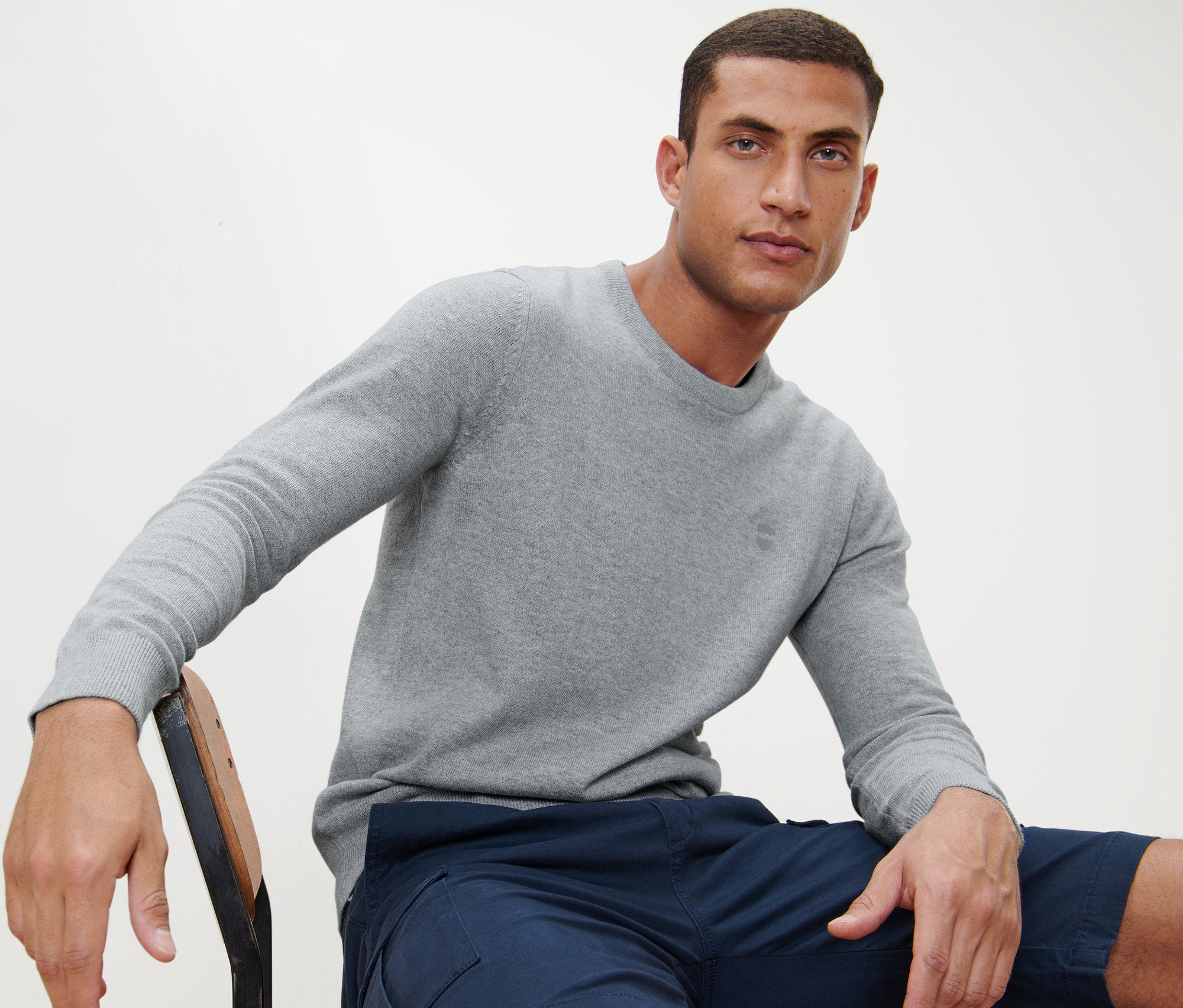 Mann sitzt auf einem Stuhl und trägt einen grauen Pullover mit Rundhalsausschnitt.