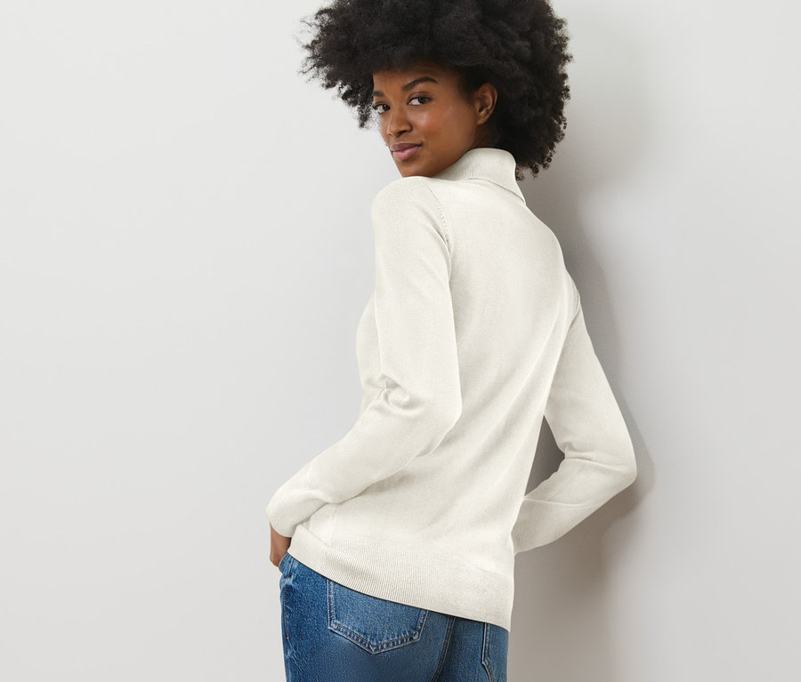 Frau blickt über die Schulter und trägt einen cremeweißen Feinstrick-Rollkragenpullover und Jeans.