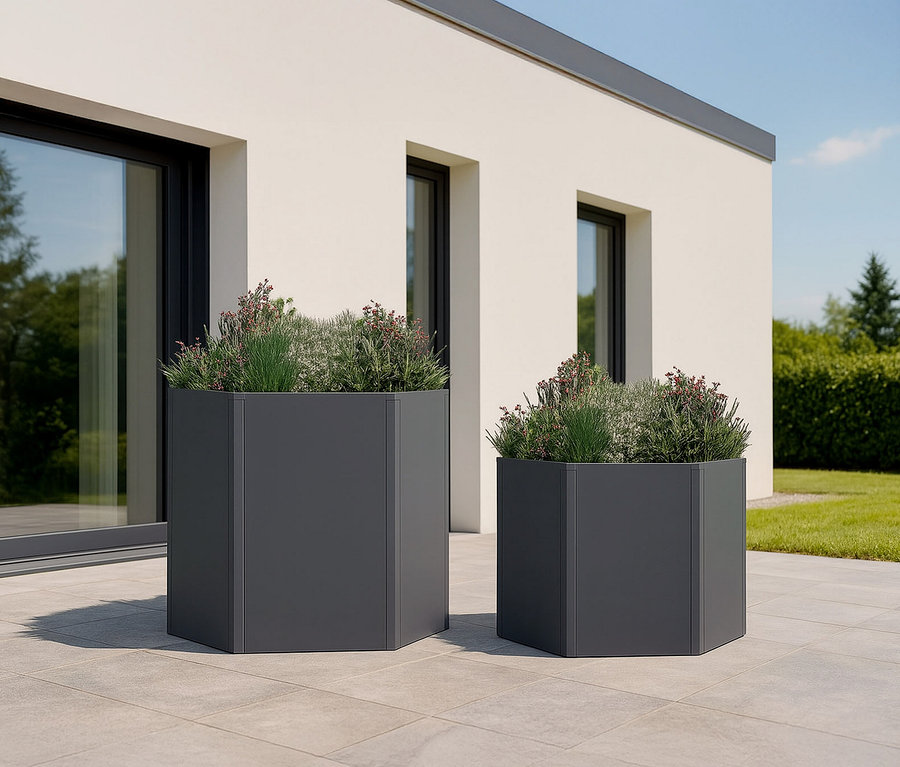 Zwei sechseckige, anthrazitfarbene Pflanzgefäße unterschiedlicher Größe stehen auf einer Terrasse vor einem Haus. Die Gefäße sind mit verschiedenen Pflanzen bepflanzt.