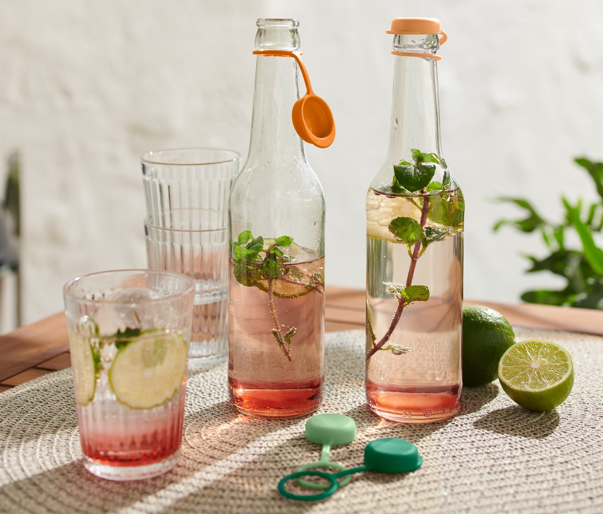 Zwei Glasflaschen mit Getränk, zwei Gläser, Limetten und 4 Flaschenverschlüsse auf einem Tisch.