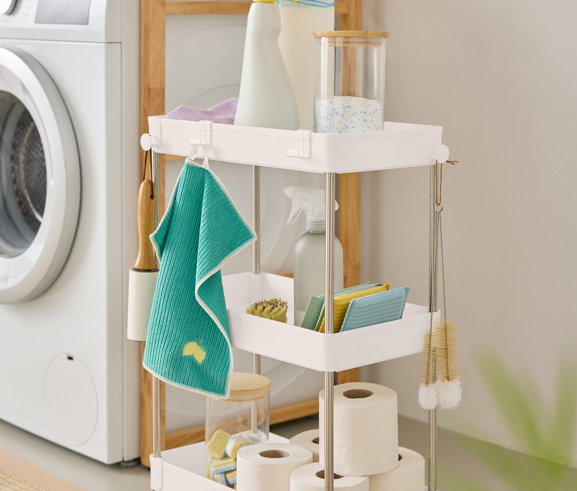 Ein dreistöckiger, weißer Rollwagen mit Reinigungsmitteln steht neben einer Waschmaschine.