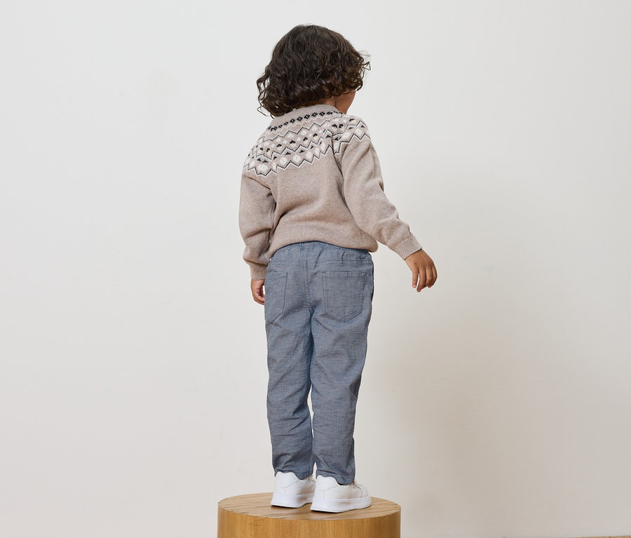 Junge steht mit dem Rücken zum Betrachter, trägt einen beigen Pullover mit Muster, blaue Hosen und weiße Schuhe.