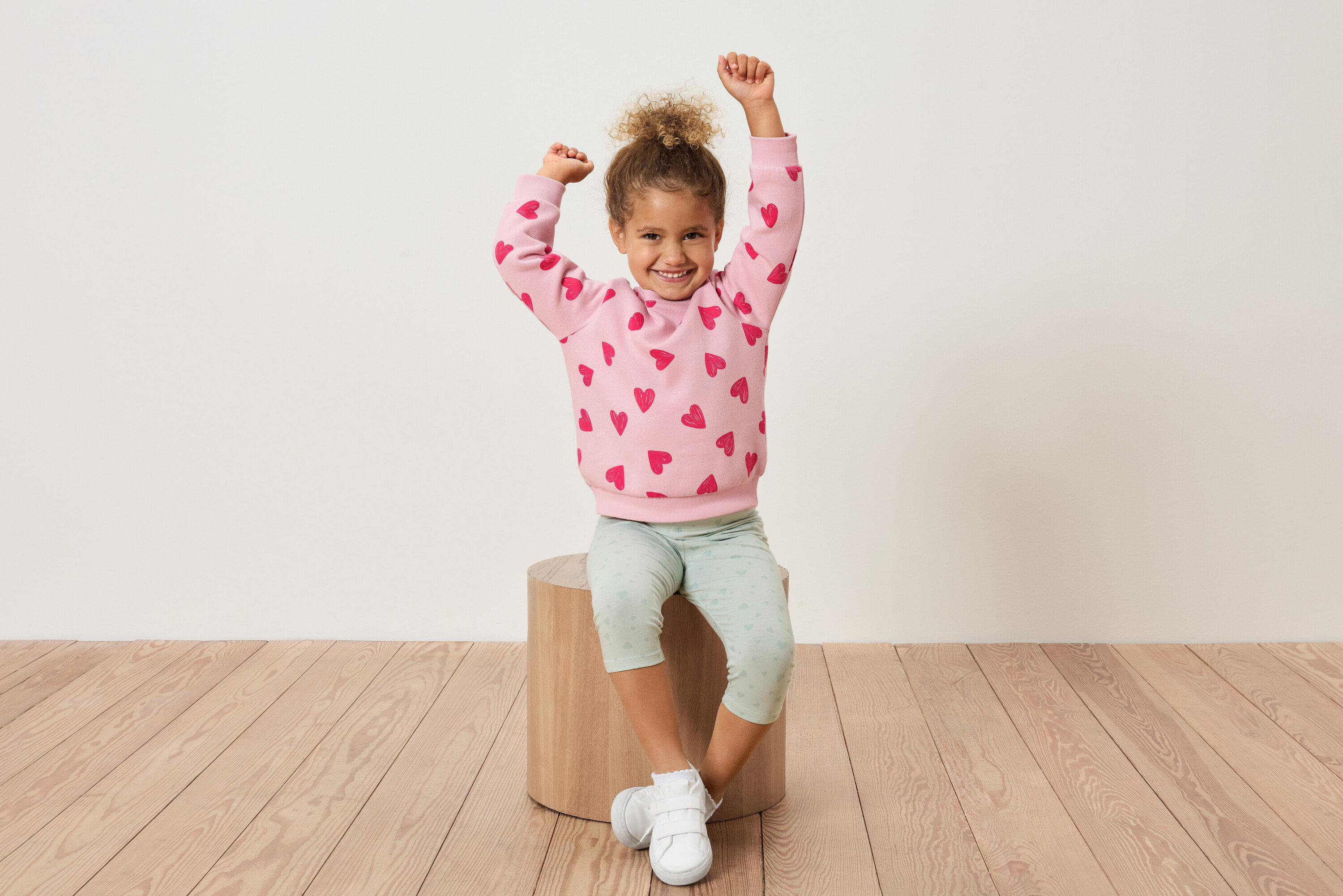 Ein junges Mädchen sitzt auf einem Holzhocker, die Arme hochgestreckt, und trägt ein rosa Sweatshirt mit roten Herzen und hellgrüne Leggings.