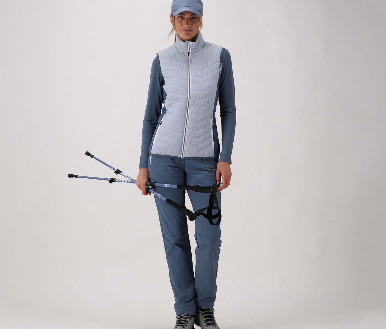 Frau in blauer Sportbekleidung mit CMP Damen-Hybridweste hält Wanderstöcke vor hellem Hintergrund.