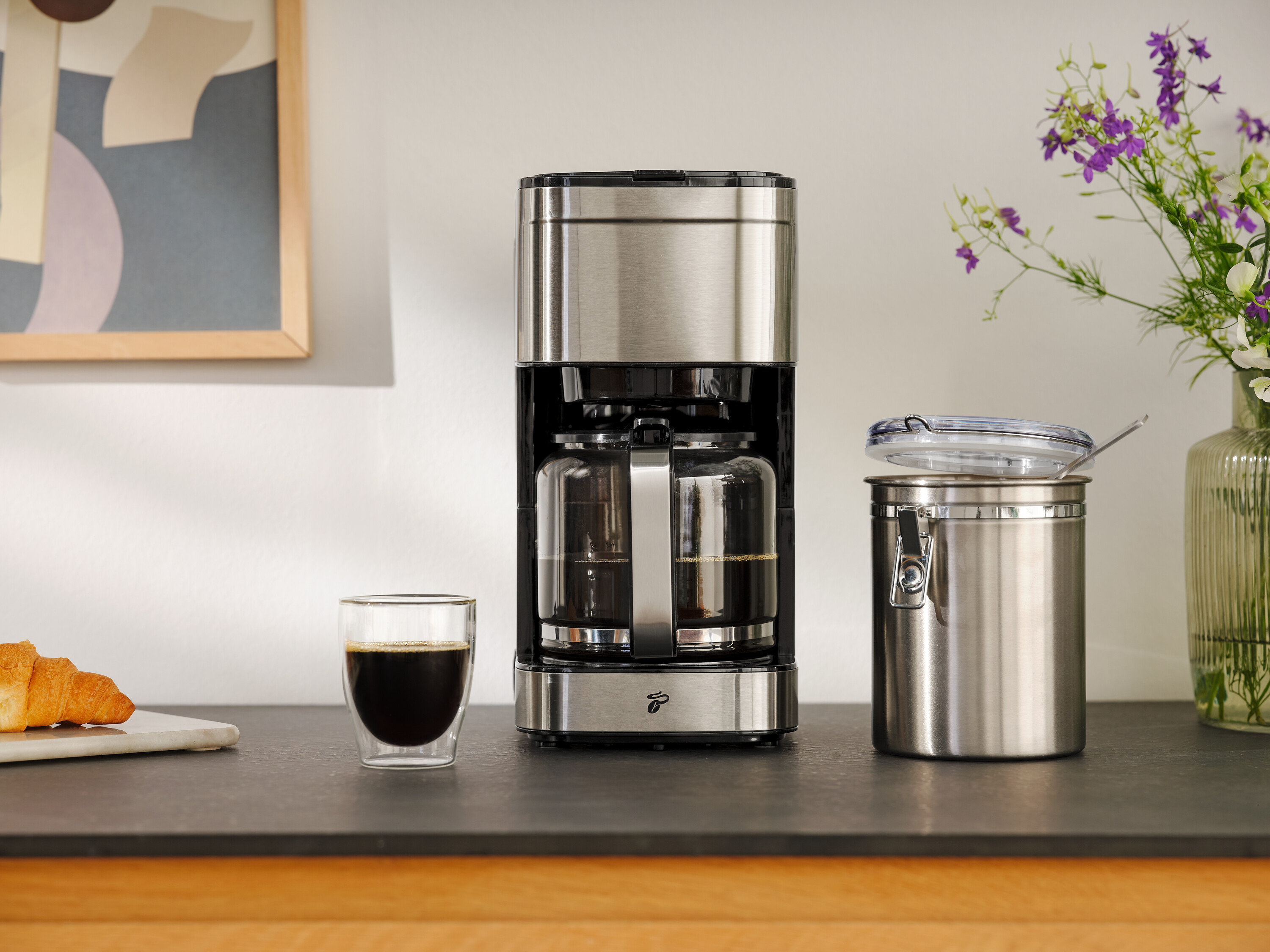 Kaffeemaschine neben einem Glas Kaffee, einem Croissant und einem Vorratsbehälter.