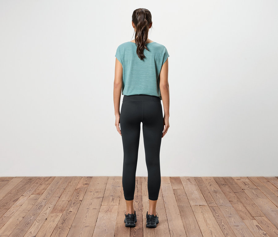 Rückansicht einer Frau mit Pferdeschwanz, die ein blaugrünes Oberteil, schwarze Leggings und schwarze Turnschuhe auf einem Holzboden trägt.